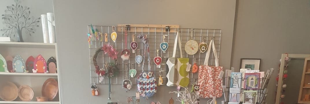 The craft wall: felt hearts and a knitted owl on a wire grid, printed tote bags, a jewellery stand, a card spinner, turned wooden bowls and painted wooden fairy doors on shelves.